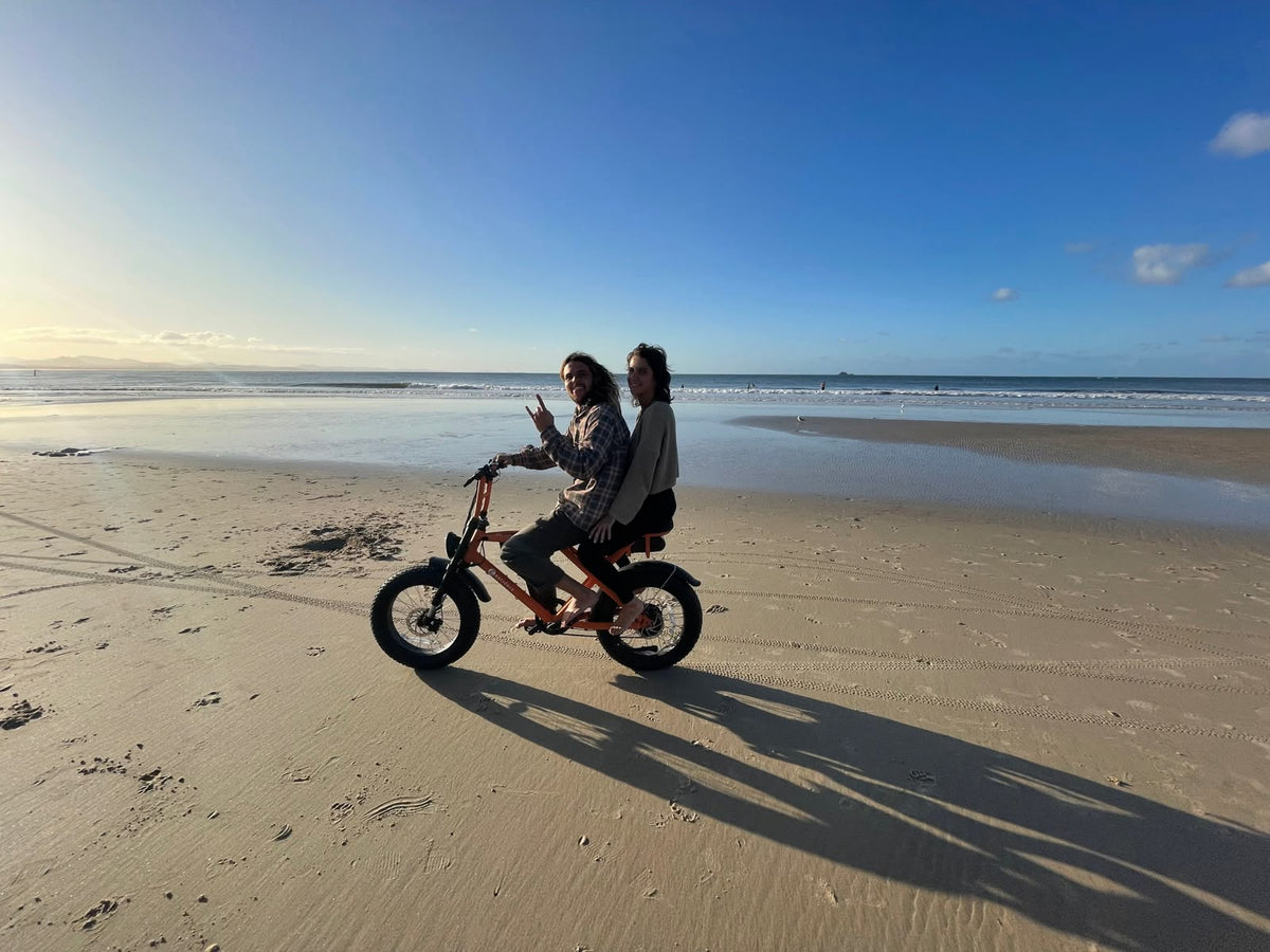 Two people on an orange electric bike beside the ocean